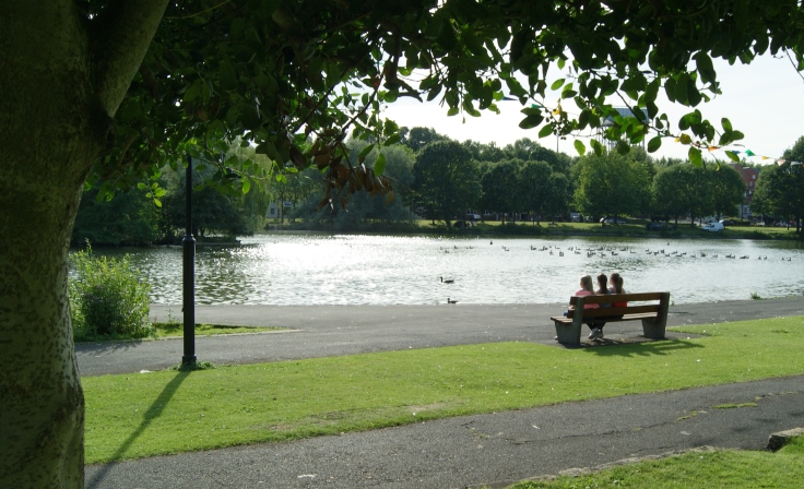 A group of people sat on a bench in front of a boating lake.